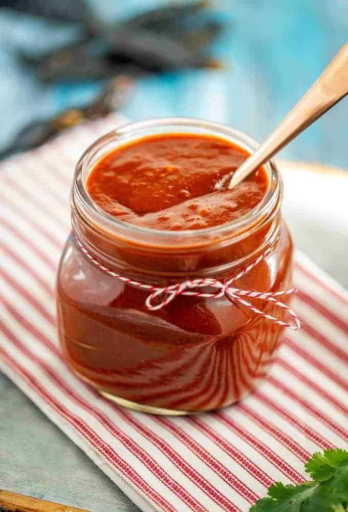 Photo of Keto Enchilada Sauce in a glass jar with a spoon in it sitting on a red and white napkin.