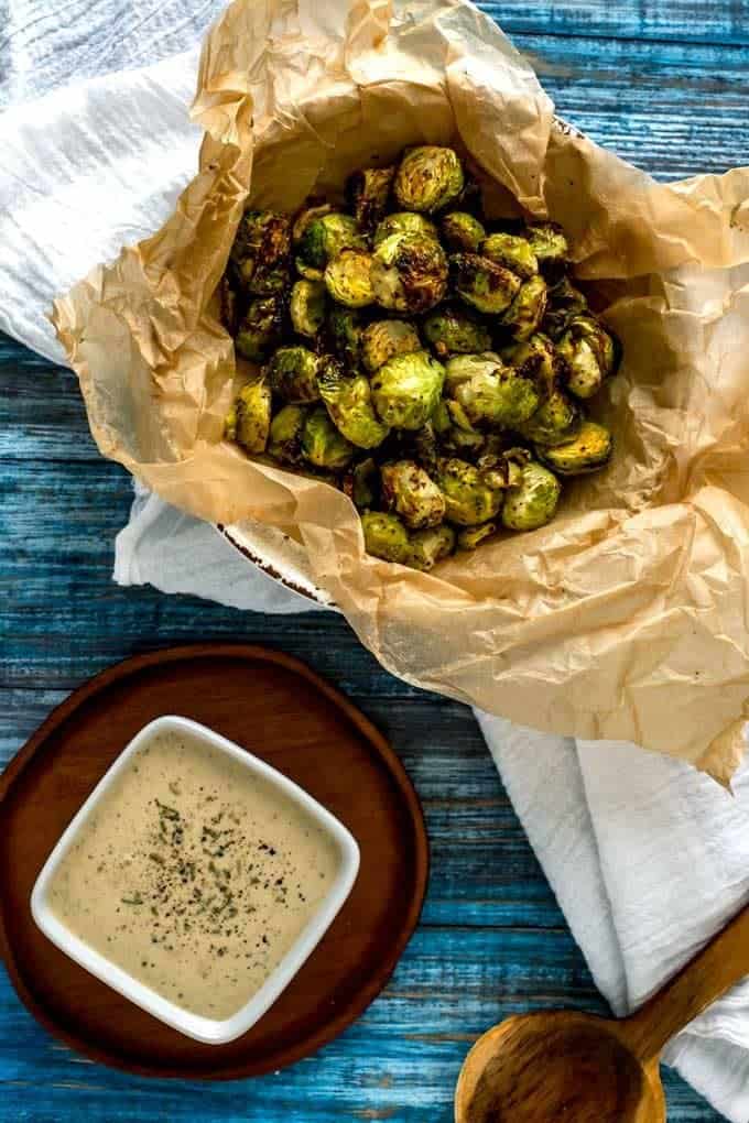 Overhead photo of Keto Brussels Sprouts in a parchment lined bowl on a blue background with a dipping sauce sitting beside it.