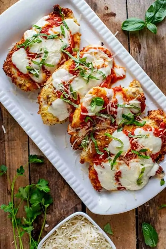 White platter of Keto Chicken Parmesan garnished with basil with parsley, basil, and Parmesan cheese around it.
