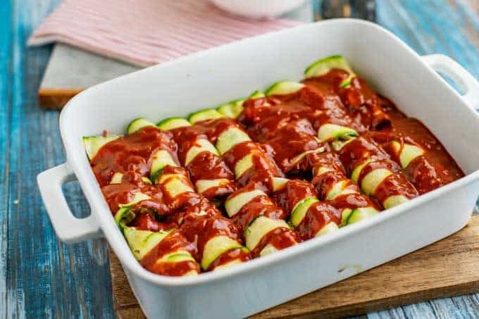 Photo of Zucchini Enchiladas topped with enchilada sauce in a white casserole dish.