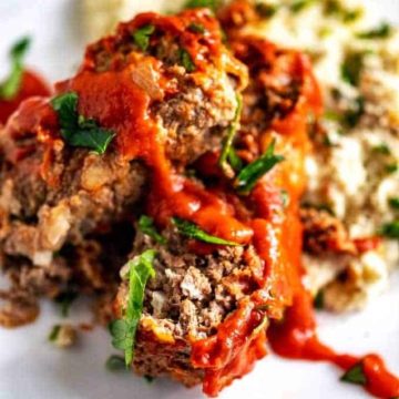 Photo of Keto Meatloaf sitting on mashed cauliflower on a white plate garnished with basil.