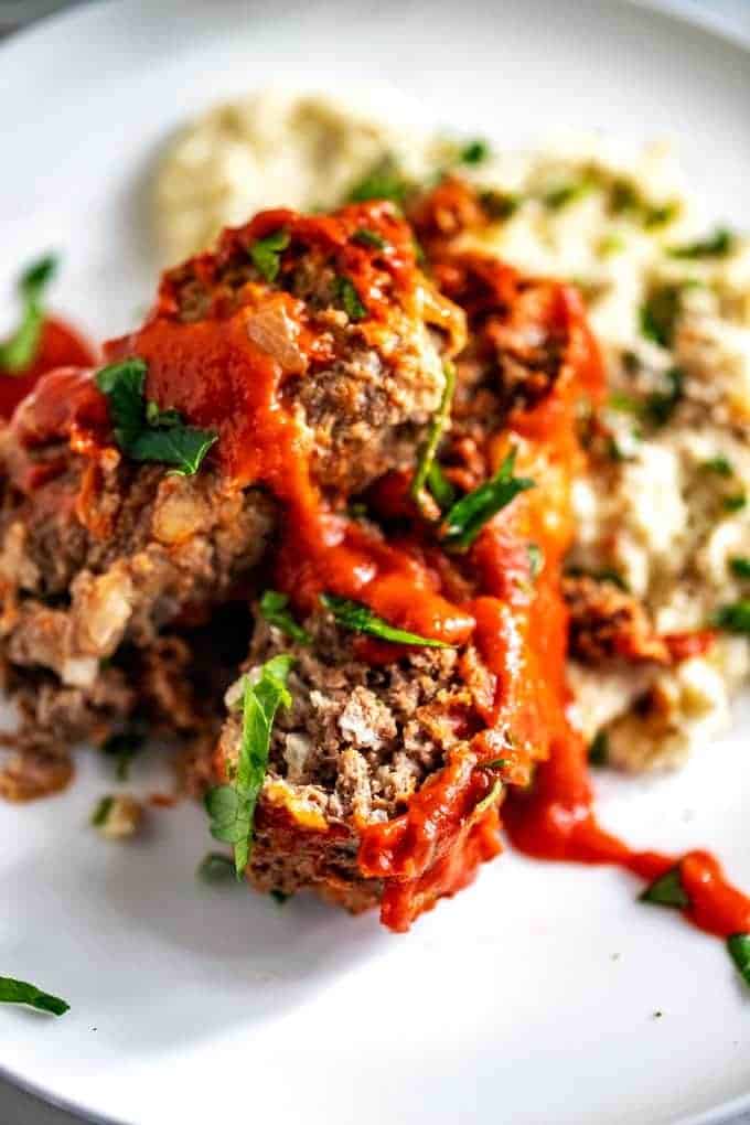 Photo of Keto Meatloaf sitting on mashed cauliflower on a white plate garnished with basil.