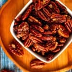 Overhead photo of Keto Pecans in a small white bowl sitting on a wooden plate against a blue background.
