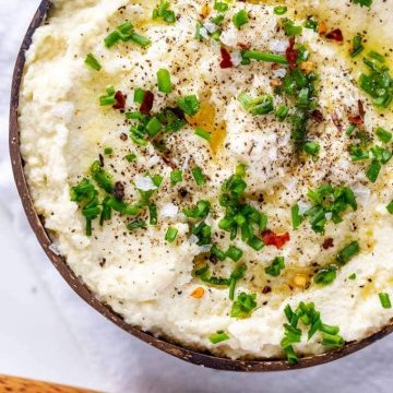 Overhead photo of low carb cauliflower mash garnished with chives, flakey salt, fresh ground pepper and crushed red pepper in a brown wooden bowl with a wooden spoon next to it.
