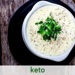 Overhead photo of Alfredo Sauce in a small white crock garnished with basil and pepper with the text below "Keto Alfredo Sauce"