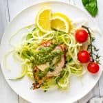 Photo of Keto Pesto Chicken sitting on a bed of zucchini noodles on a white plate with lemon and cherry tomatoes on the plate.
