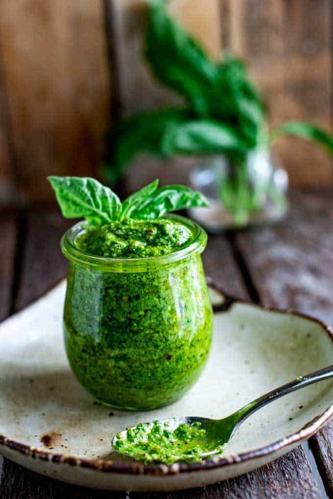 Side photo of low carb pesto in a glass jar sitting on a plate with a spoon by it on a dark background.