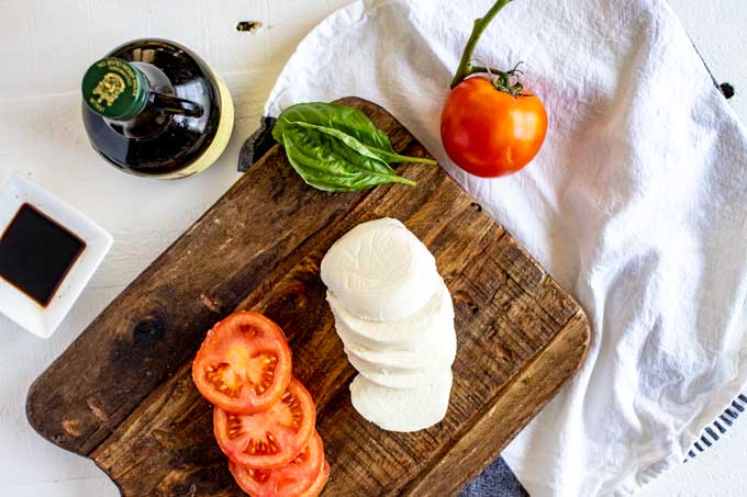 Overhead photo of ingredients for keto caprese salad:  sliced tomato, mozzarella, basil, olive oil, and balsamic vinegar.