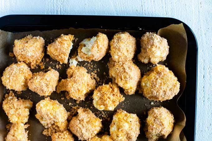 Photo of a baking sheet lined with parchment paper with cauliflower florets that have been breaded with an almond flour/Parmesan cheese mixture.