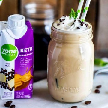 Photo of a Keto Iced Latte in a jar with two straws and a ZonePerfect Keto shake sitting next to it on a white cutting board agains a dark wooden background.