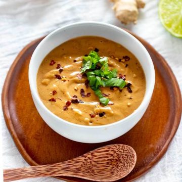 Photo of Keto Peanut Sauce in a white bowl garnished with Thai Basil and crushed red pepper flakes sitting on a wooden plate with a wooden spoon above it.