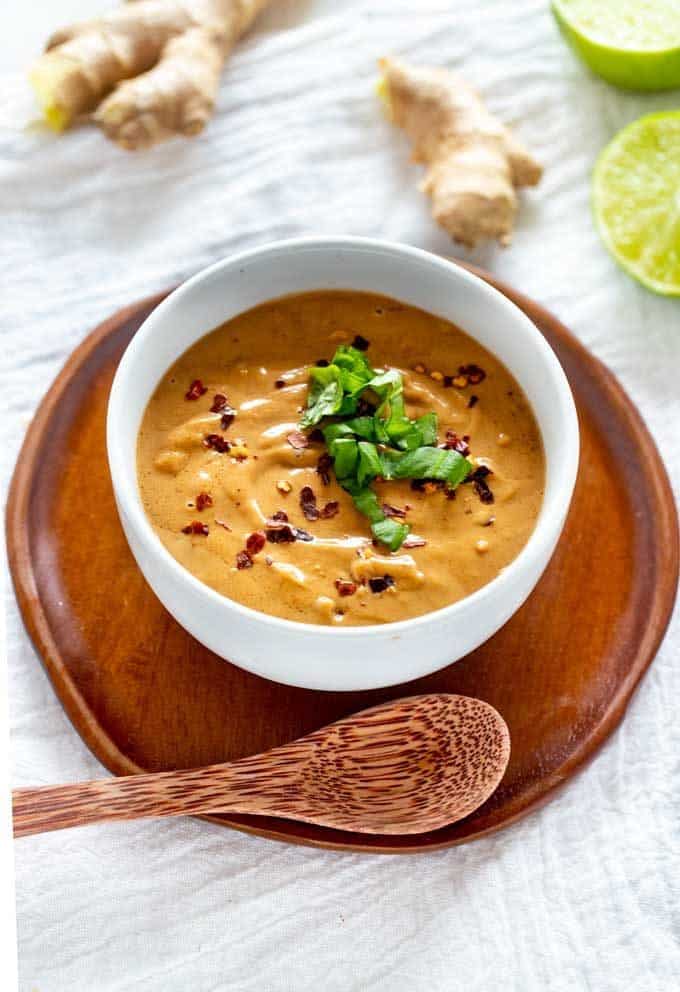 Photo of Keto Peanut Sauce in a white bowl garnished with Thai Basil and crushed red pepper flakes sitting on a wooden plate with a wooden spoon above it.