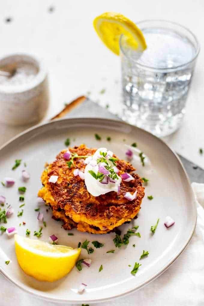 ¾ Overhead Photo of Zucchini Fritters Keto Style garnished with parsley and red onion with a lemon sitting next to it and a glass of water and salt cellar sitting behind it.