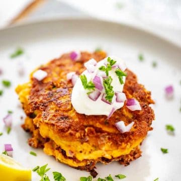 Close up photo of Keto Zucchini Fritters with a dollop of sour cream on top garnished with parsley and red onion sitting on a light grey plate with a lemon wedge next to it.