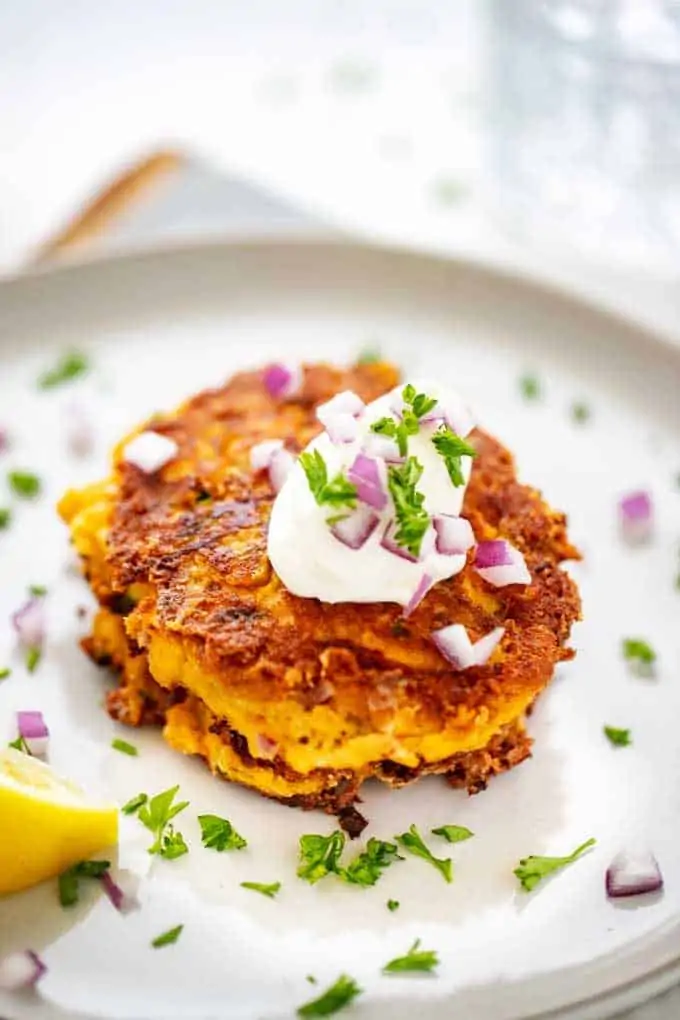 Close up photo of Keto Zucchini Fritters with a dollop of sour cream on top garnished with parsley and red onion sitting on a light grey plate with a lemon wedge next to it.