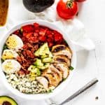 Overhead photo of a Low Carb Cobb Salad in a white bowl on a white background surrounded by avocado, tomato, and dressing.