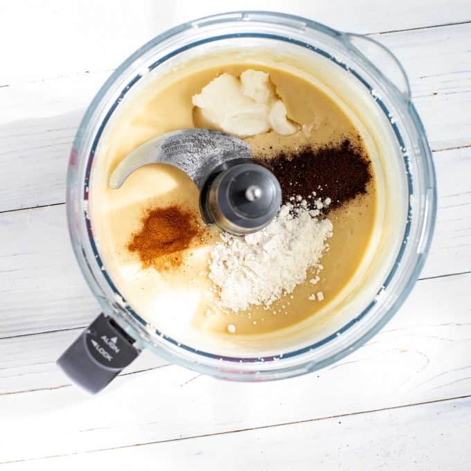 Photo of vanilla powder, cinnamon, Swerve, and coconut oil being added to a food processor with macadamia nut butter in it.