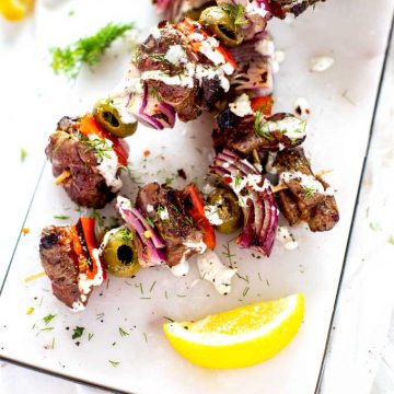 Overhead photo of Keto Lamb Kebabs on a white marble cutting board drizzles with a cream sauce and garnished with dill.