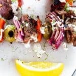 Close up photo of grilled lamb kebabs drizzled with sauce on a white plate with a lemon.