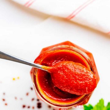 Overheat photo os Low Carb Ketchup in a jar with a spoon of ketchup resting on it sitting on a white background with parsley and red pepper flakes scattered.