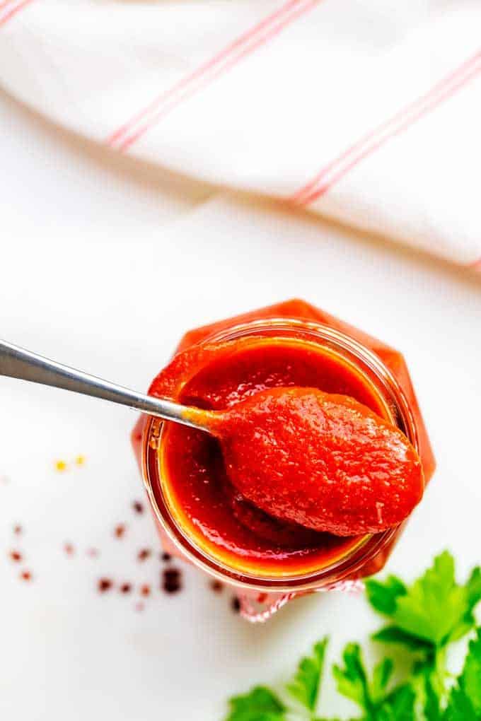 Overheat photo os Low Carb Ketchup in a jar with a spoon of ketchup resting on it sitting on a white background with parsley and red pepper flakes scattered.
