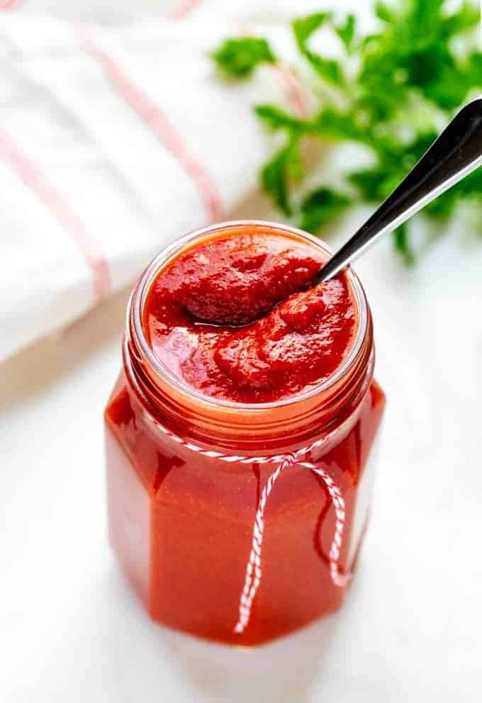 Photo of Low Carb Ketchup in a glass jar with a spoon in it sitting on a white background with a red and white napkin behind it.
