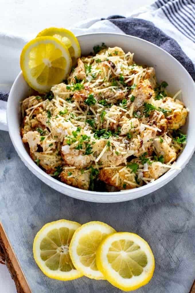 Side overhead photo of Low Carb Roasted Cauliflower in a white bowl sitting a a grey weathered cutting board garnished with lemons and parsley.