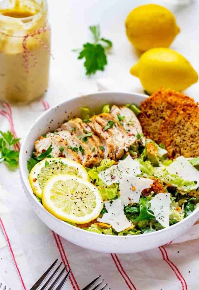 Photo of a Low Carb Caesar Salad in a white bowl with two lemons and a jar of homemade caesar dressing sitting behind it.