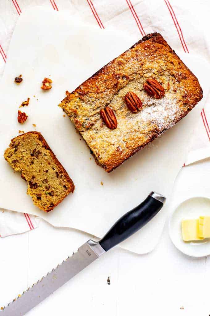 Photo of Keto Zucchini Bread with pecans on top sitting on a marble cutting board with a knife next to it and a slice cut out of it.