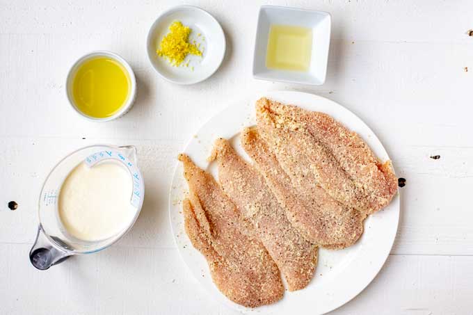 Photo of chicken that has been coated in a Parmesan/Almond flour mixture with small prep bowls containing lemon juice, lemon zest, oil, and heavy cream next to it.