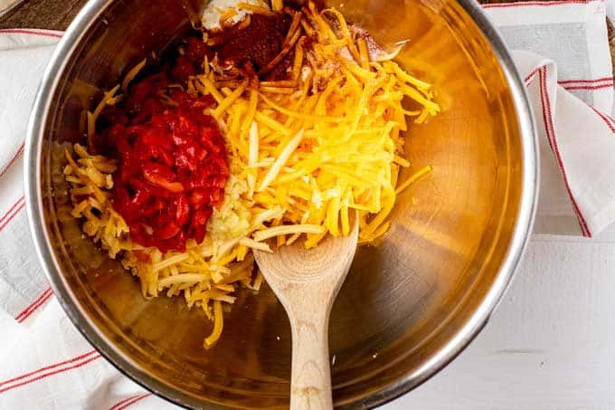 Photo of mayonnaise, cream cheese, sharp cheddar cheese, smoked gouda, garlic, pimentos, salt, smoked paprika, and cayenne pepper in a large bowl being mixed together.