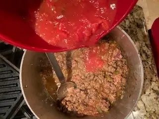 Photo of tomatoes being added to a dutch oven with browned ground beef.