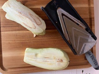 Photo of eggplant being sliced thin with a madoline slicer.