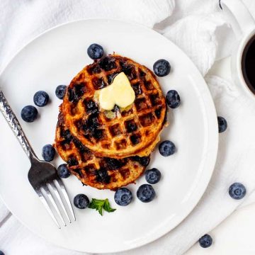 Overhead photo of two Keto Blueberry Chaffles on a white plate surrounded by blueberries.