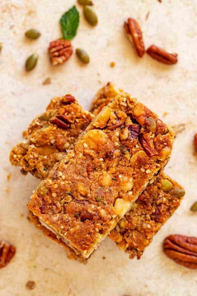 Overhead photo of three stacked Keto Granola Bars on a travertine background with nuts and seeds scattered around.