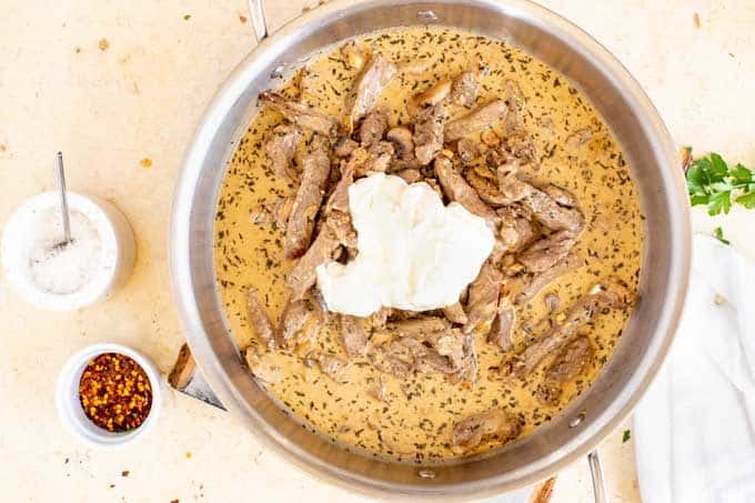 Sour cream being added to a large skillet with a creamy sauce, steak, and mushrooms.