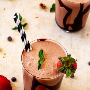 Photo of two glasses of a Keto Chocolate Smoothie garnished with strawberries and mint, with strawberries and chocolate chips scattered around them.