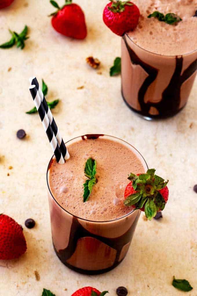 Photo of two glasses of a Keto Chocolate Smoothie garnished with strawberries and mint, with strawberries and chocolate chips scattered around them.
