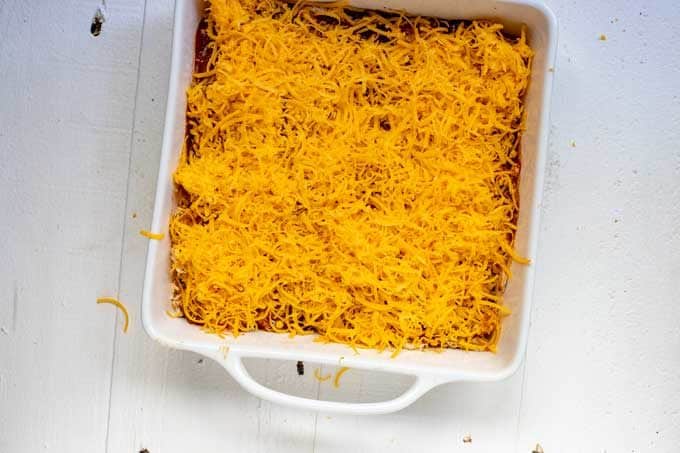 Photo of a white square casserole dish with cheddar cheese sprinkled on top of the filling.