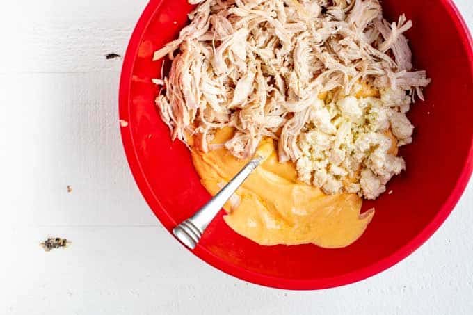 Red bowl with shredded chicken being mixed in with a blend of cream cheese, mayonnaise, ranch dressing, and buffalo sauce.