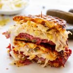 A stacked keto Reuben sandwich filled with corned beef, sauerkraut, melted cheese, pickles, and dressing is held together with a toothpick. The sandwich is cut in half and sits on a white surface with pickles in the background.
