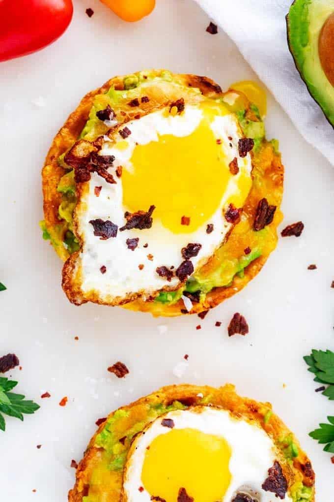 Overhead photo of two Keto Avocado Toasts on a white background with fried egg, cheese, and bacon.
