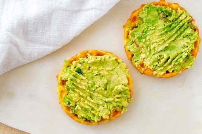 A Keto Chaffle with mashed avocado  on it on a white cutting board.