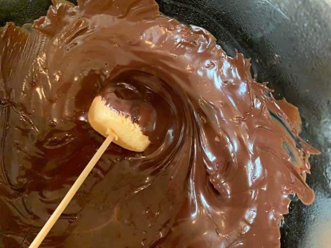 Photo of a peanut butter ball on a wooden skewer being dipped into chocolate.