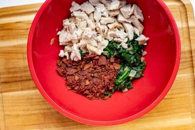 A red bowl with chopped cook chicken, crumbled cooked bacon, and sauteed spinach.