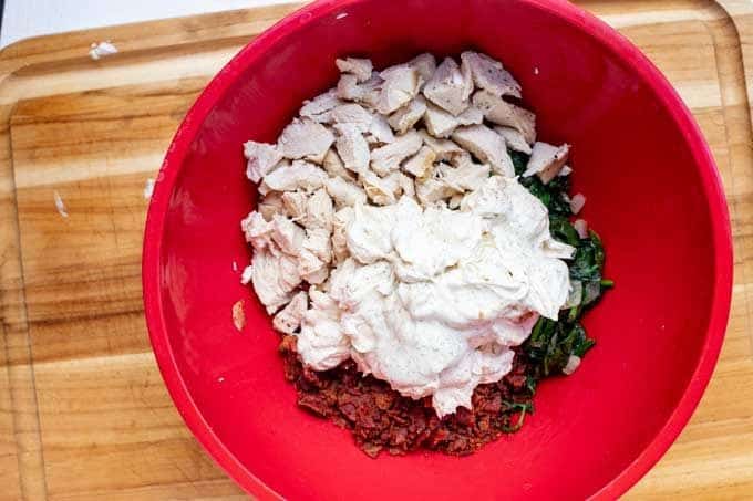 A red bowl with chopped cook chicken, crumbled cooked bacon, sauteed spinach and ranch dressing.