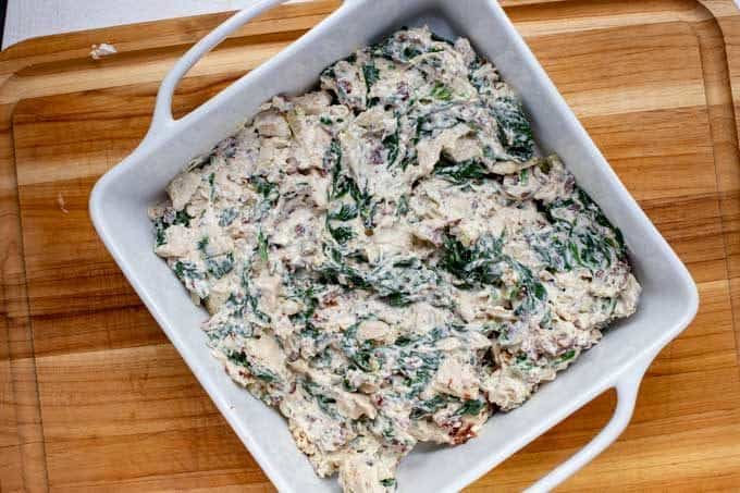 A white casserole dish with chopped cook chicken, crumbled cooked bacon, sauteed spinach and ranch dressing that have been mixed together.