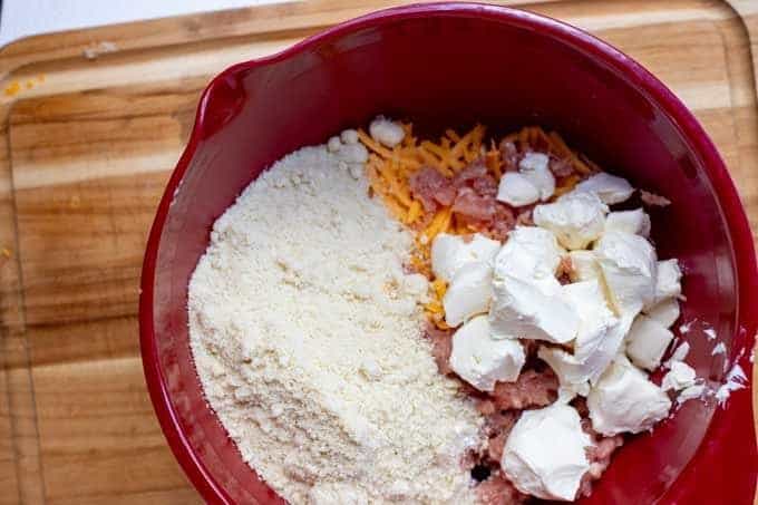 Photo of breakfast sausage, cream cheese, almond flour, shredded cheddar, and seasonings in a red bowl sitting on a cutting board.