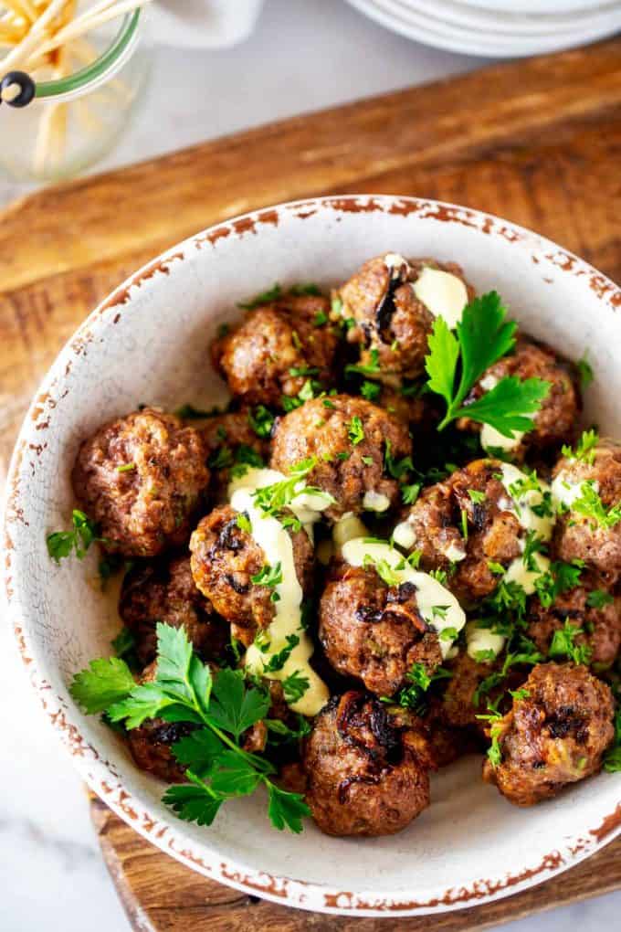 Photo of air fryer caramelized onion meatballs in a rustic white bowl with a mayo mustard drizzle over them garnished with parsley.