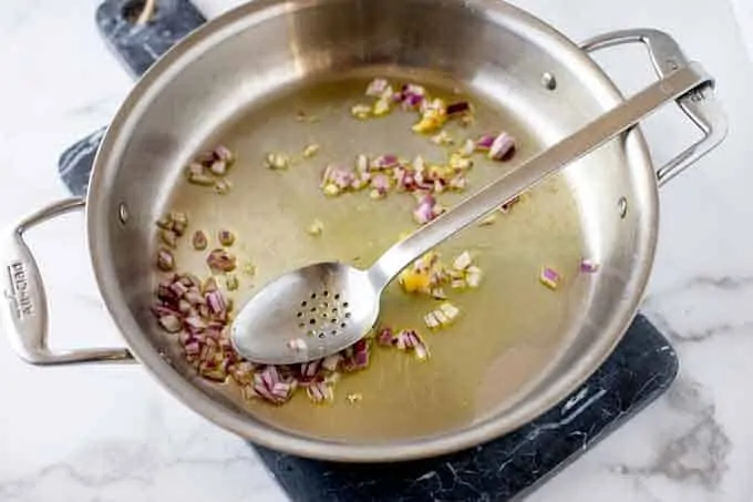 Photo of a skillet with onion, oil, and garlic with a metal spoon.